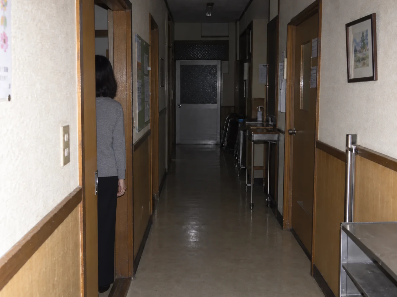 集会所の廊下。左の戸口に、灰色の上着と黒い下衣の人物が室内へ向いた後ろ姿で立っている。顔は写っていない。